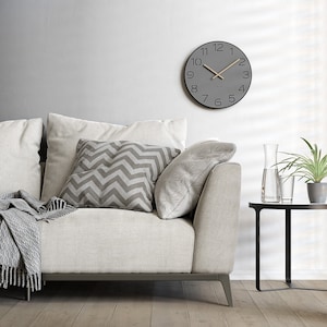 Könnte beinhalten: Ein modernes Wohnzimmer mit einer grauen Wanduhr mit schwarzen Ziffern und Zeigern. Ein helles Sofa ist mit grau-weiß gemusterten Kissen und einer passenden Decke ausgestattet. Ein kleiner schwarzer Beistelltisch trägt eine Glaskaraffe und eine Topfpflanze.