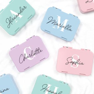 Könnte beinhalten: Eine Sammlung von personalisierten Mint-Dosen in Pastellfarben. Jede Dose hat eine andere Farbe, darunter Hellblau, Rosa und Lavendel. Die Dosen sind mit Namen in Schreibschrift beschriftet, mit einem großen Anfangsbuchstaben.