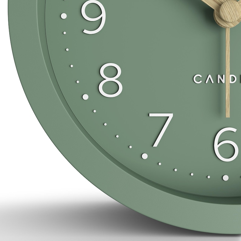 May include: Close-up of a sage green analog clock with white numerals and a light wood-colored clock hand. The clock face has the word "CANDI" printed in white. The clock is a modern design.