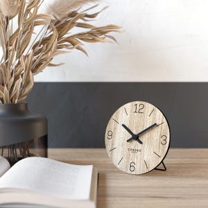 Può includere: Un orologio rotondo in legno con lancette e numeri neri. Il quadrante dell'orologio ha un effetto venatura del legno chiaro e un supporto in metallo nero. Le parole "CANDER BERLIN" sono stampate sul quadrante. L'orologio è su una superficie di legno.
