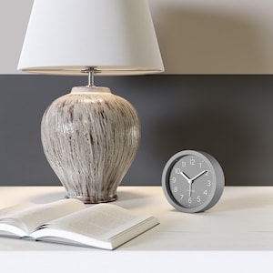Può includere: Una composizione da comodino con una sveglia analogica grigia, una lampada in ceramica con paralume bianco e un libro aperto. La lampada ha una base strutturata bianco sporco con accenti marroni. L'orologio mostra l'ora con lancette e numeri bianchi.