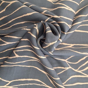 May include: A close-up shot of a folded fabric. The fabric is a blue-grey colour with a repeating pattern of off-white, wavy lines. The material has a soft texture and a subtle sheen, possibly silk or linen.