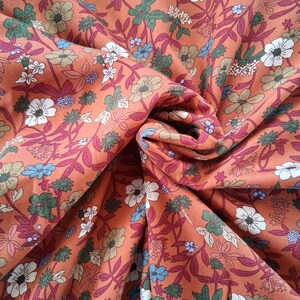 May include: Close-up of a floral fabric with a warm orange background. The fabric features a repeating pattern of white, blue, and green flowers with burgundy stems and leaves. The fabric is folded, creating shadows and depth.