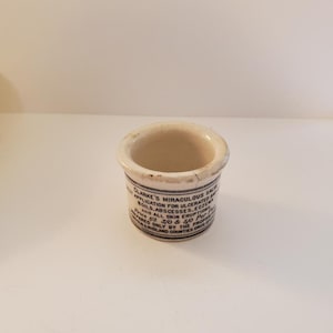 May include: A small white ceramic jar with blue lettering that reads "Clarke's Miraculous Salve Application for Ulcerated Boils, Abscesses, Eczema and All Skin Eruptions! Price 1/3 3/0 & 5/0 Per Pot Prepared Only by the Proprietors Midland Counties Drug Stores"