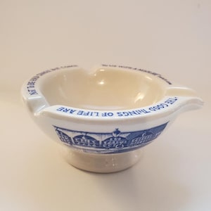 May include: White ceramic ashtray with a blue and white design. The ashtray has a wide, shallow bowl and a handle on one side. The text "The good things of life are not to be had without some courage" is printed around the rim of the bowl.
