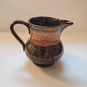 May include: A small, brown ceramic creamer with a gold and orange design. The creamer has a handle and a spout.