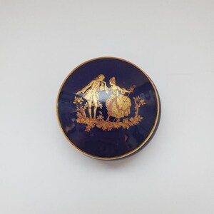 May include: A small, round, blue and gold trinket box with a lid. The box features a gold design of a couple in a garden setting.