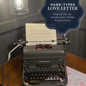 May include: A black Remington typewriter with a typed letter in the carriage. The image text reads "HAND-TYPED LOVE LETTER Typed on an Authentic 1940s Typewriter". The typewriter is on a wooden table with floral patterned paper.