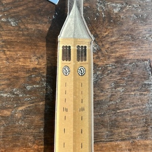 May include: A clear acrylic ornament shaped like a tall building with a clock face on each side. The building is painted in shades of brown and yellow with black accents. The ornament is a representation of a famous landmark.