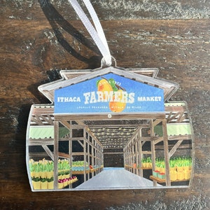 May include: A clear plastic ornament shaped like a building with a sign that reads "Ithaca Farmers Market, Since 1973, Locally Produced Within 30 Miles". The ornament depicts the interior of a market with produce and flowers on display.