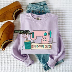Peut inclure: Un sweat-shirt lilas avec un motif de machine à coudre rose, des ciseaux et une bobine de fil turquoise. Le texte "Proverbes 31:13" est en dessous. Le sweat-shirt est associé à un jean bleu et des chaussures marron.