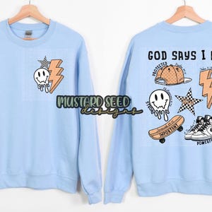May include: Light blue sweatshirt with a front graphic of a smiley face, lightning bolt, and checkered star. The back features the text "GOD SAYS I AM" with additional graphics including a skateboard, sneakers, and a baseball cap. The sweatshirt is displayed on a wooden hanger.