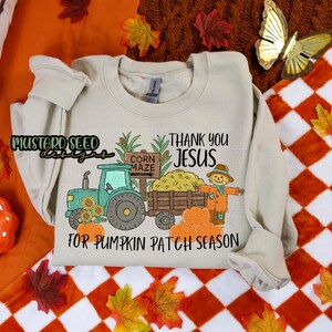 Op de afbeelding: Crèmekleurige sweatshirt met een herfstthema. Het ontwerp bevat een groene tractor, zonnebloemen, pompoenen en een vogelverschrikker. De tekst luidt "Thank You Jesus" en "For Pumpkin Patch Season."