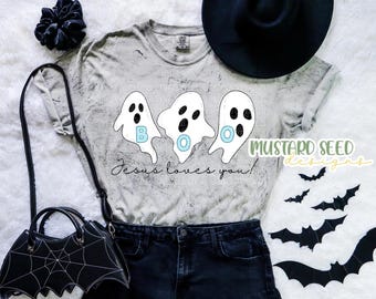 Könnte beinhalten: Hellgraues T-Shirt mit einem "BOO" Geister-Design und dem Text "Jesus loves you!". Das Shirt wird mit schwarzen Jeansshorts, einer fledermausförmigen Handtasche und einem schwarzen Hut kombiniert, was ein Halloween-Thema ergibt.