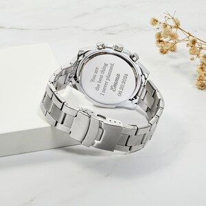 Puede incluir: Reloj plateado con una correa de acero inoxidable. La parte posterior del reloj tiene un mensaje grabado que dice: "You are the best thing I never planned. Emma 06.20.2024"