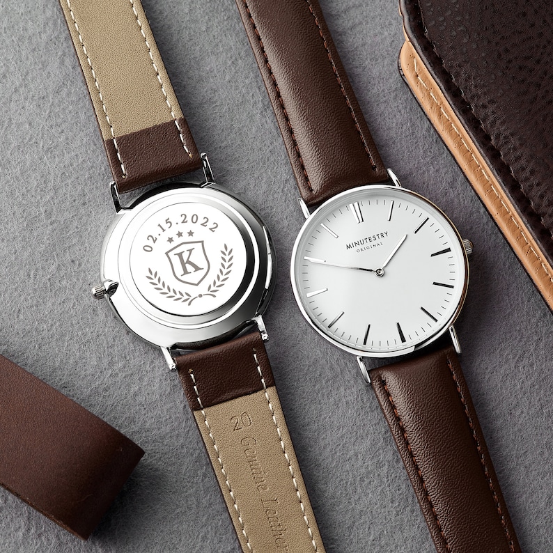 Two brown leather strap watches, one showing the engraved back with a personalized design featuring the date 02.15.2022 and a crest with the letter K, while the other watch displays the minimalist white dial from the front