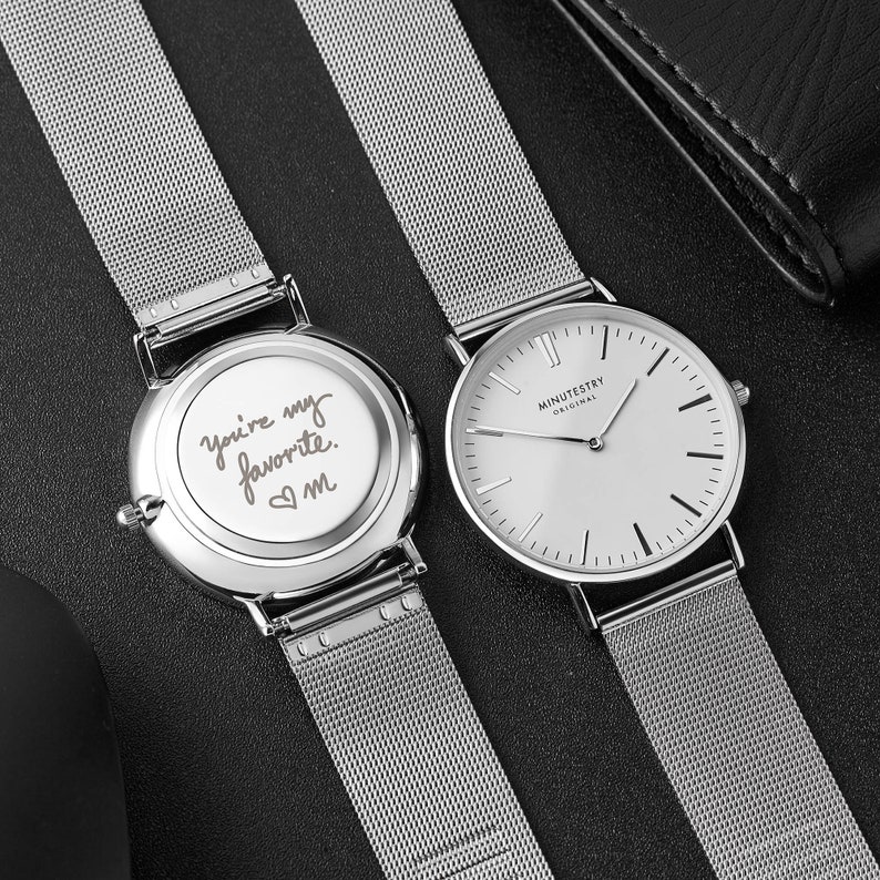 A watch with a silver mesh strap and minimalist white dial, personalized with an engraved handwritten message on the back.