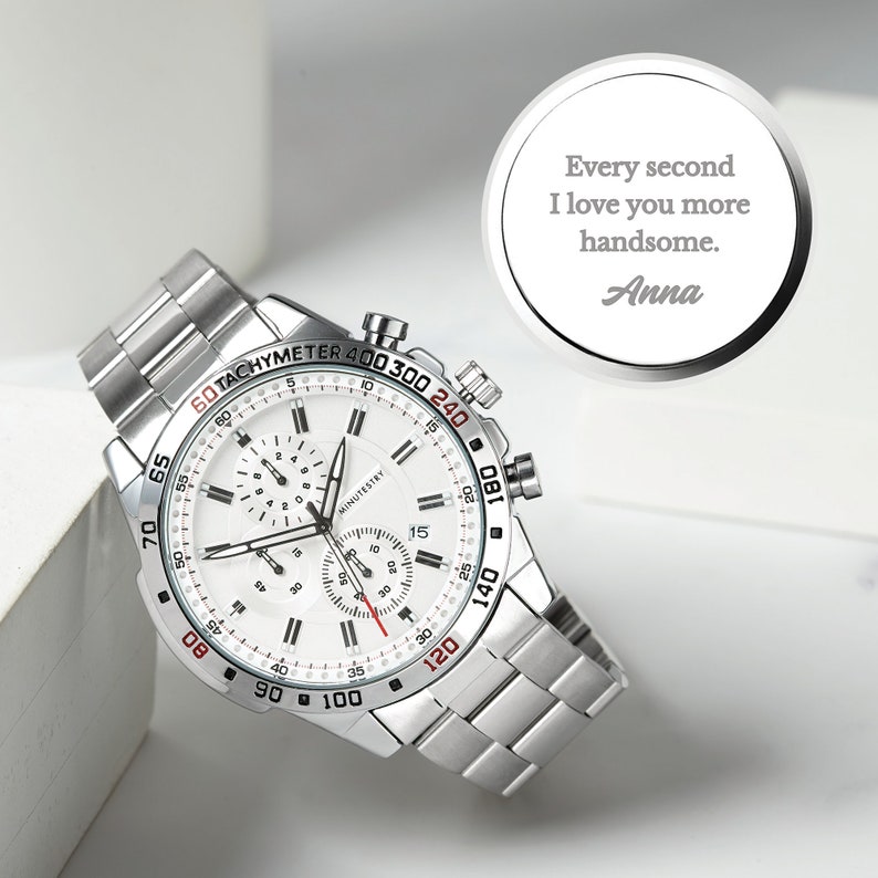 May include: A silver chronograph wristwatch with a white face and red accents. The watch has a tachymeter scale around the outer edge of the face. The watch is on a silver metal band. The watch face has the text "MINUTESTRY" printed on it. The watch is accompanied by a silver circular tag with the text "Every second I love you more handsome. Anna".