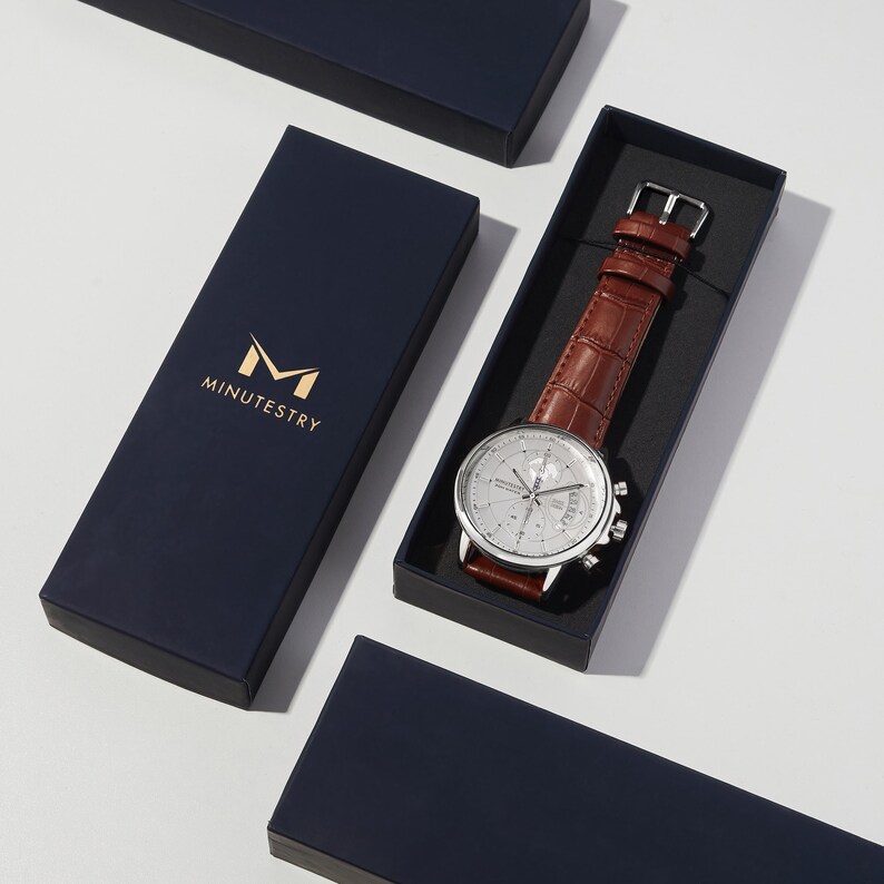 May include: A silver watch with a white face and brown leather band in a navy blue box with the logo "M MINUTESTRY".