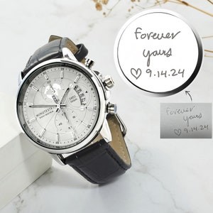 May include: A silver watch with a black leather band. The watch face has a white background with black markings and a date window. The watch has a chronograph function. The watch is on a white surface with a silver disc that says "forever yours 9.14.24" written on it.