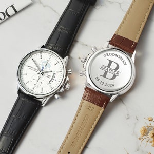 May include: Two silver wristwatches with black and brown leather bands. The watch on the left has a white face with a date window and a chronograph. The watch on the right has a silver face with the text "GROOMSMAN" and "BRAYDEN" engraved on the back. The date "08.12.2024" is also engraved on the back.