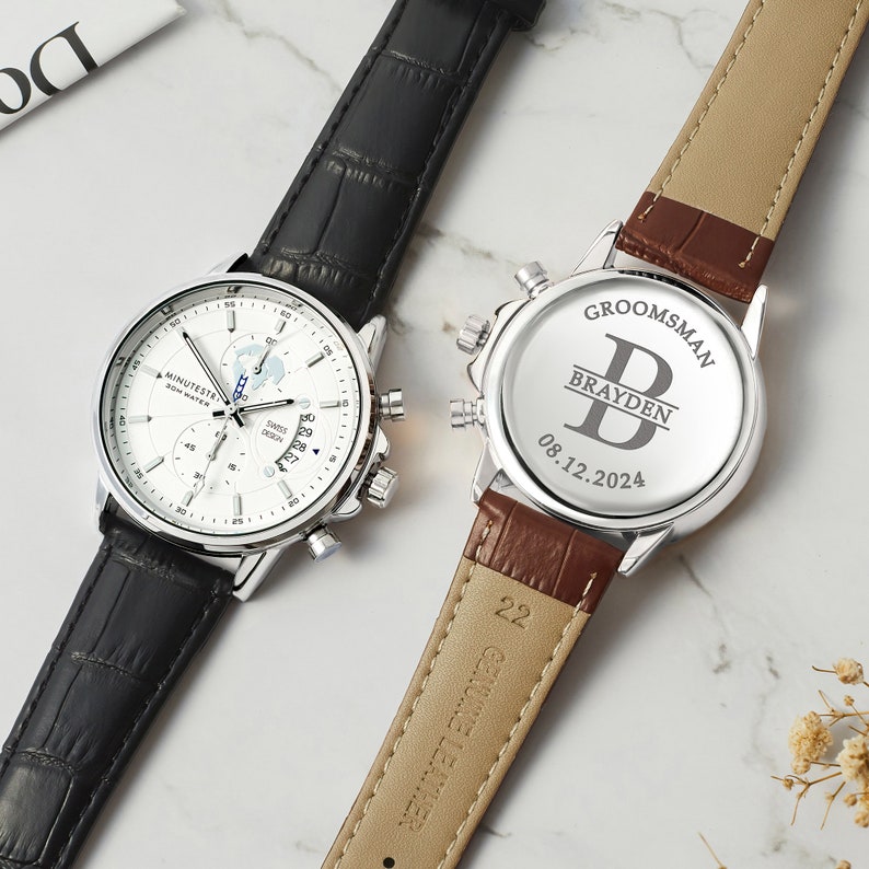 May include: Two silver wristwatches with black and brown leather bands. The watch on the left has a white face with a date window and a chronograph. The watch on the right has a silver face with the text "GROOMSMAN" and "BRAYDEN" engraved on the back. The date "08.12.2024" is also engraved on the back.