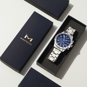 Puede incluir: Un reloj plateado con una esfera azul y una correa de cuero negro. El reloj tiene una funci&oacute;n de cron&oacute;grafo y una ventana de fecha. El reloj est&aacute; en una caja negra con la palabra "MINUTESTRY" impresa en ella.