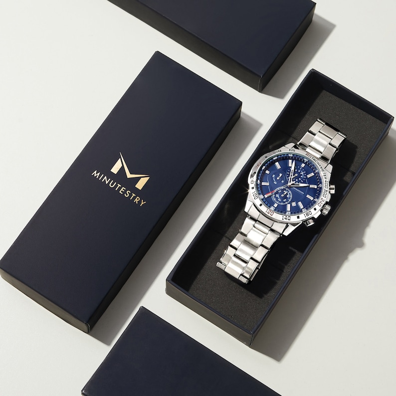 Puede incluir: Un reloj plateado con una esfera azul y una correa de cuero negro. El reloj tiene una funci&oacute;n de cron&oacute;grafo y una ventana de fecha. El reloj est&aacute; en una caja negra con la palabra "MINUTESTRY" impresa en ella.