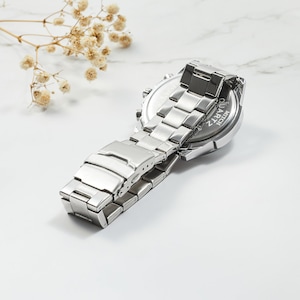Puede incluir: Reloj de pulsera plateado con una correa de acero inoxidable. La parte posterior del reloj tiene las palabras "QUARTZ" y "WR-50M" grabadas.
