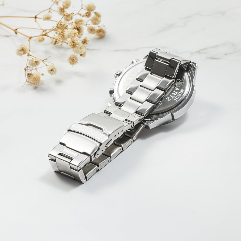 Puede incluir: Reloj de pulsera plateado con una correa de acero inoxidable. La parte posterior del reloj tiene las palabras "QUARTZ" y "WR-50M" grabadas.