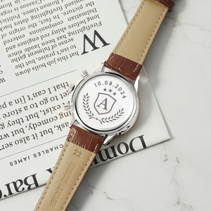 May include: A silver watch with a brown leather band. The back of the watch has an engraved design with the letter "A" inside a shield, three stars, and a laurel wreath. The date "10.08.2024" is also engraved on the back of the watch.
