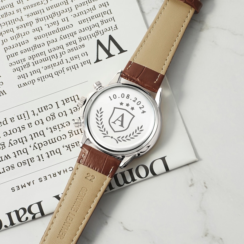 May include: A silver watch with a brown leather band. The back of the watch has an engraved design with the letter "A" inside a shield, three stars, and a laurel wreath. The date "10.08.2024" is also engraved on the back of the watch.