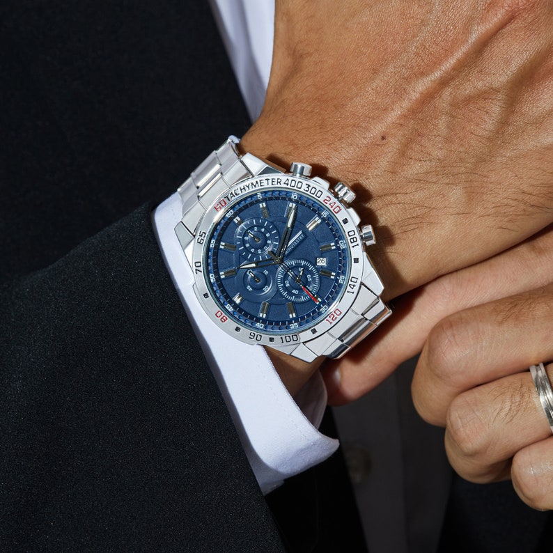 Puede incluir: Un reloj de pulsera cron&oacute;grafo plateado con una esfera azul y detalles rojos. El reloj tiene una escala de tac&oacute;metro alrededor del borde exterior de la esfera. El reloj est&aacute; en una pulsera de metal plateado.