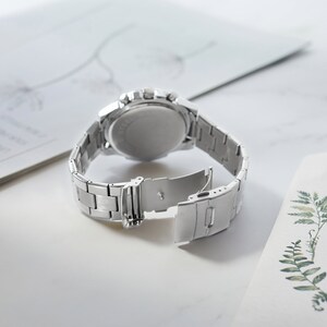 Puede incluir: Un reloj de pulsera plateado con una correa de acero inoxidable. El reloj tiene una esfera redonda y una funci&oacute;n de cron&oacute;grafo. La correa es ajustable y tiene un cierre de hebilla.