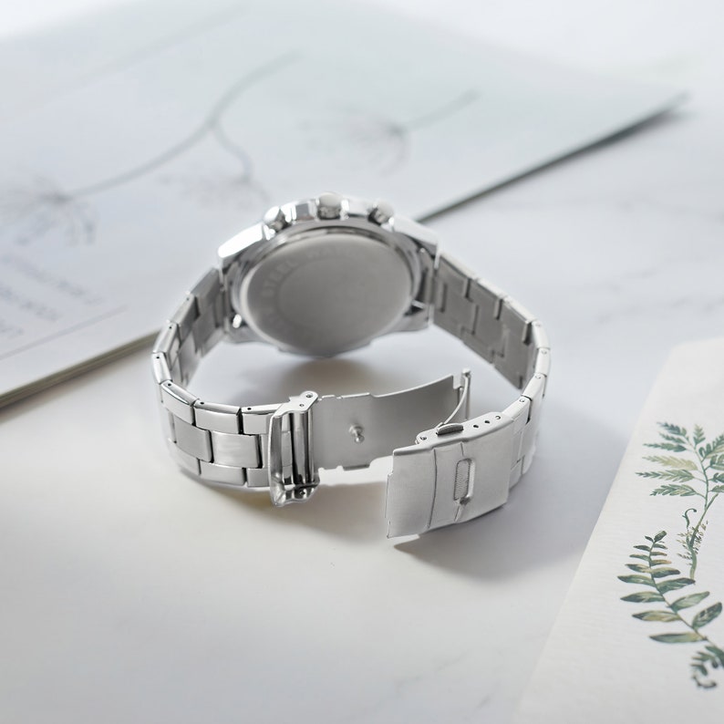 Puede incluir: Un reloj de pulsera plateado con una correa de acero inoxidable. El reloj tiene una esfera redonda y una funci&oacute;n de cron&oacute;grafo. La correa es ajustable y tiene un cierre de hebilla.