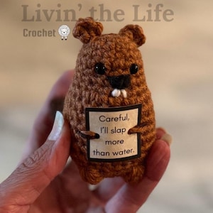 Peut inclure: Une poupée castor marron crochetée avec des yeux noirs et une dent blanche. Le castor tient une pancarte qui dit "Attention, je gifle plus que l'eau". Le texte "Livin' the Life Crochet" est en haut.