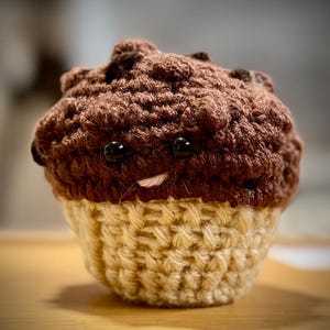 May include: A crocheted muffin with a smiling face. The top is a dark brown color, and the base is a light tan. The muffin has two black bead eyes and a small, light-colored stitched mouth.