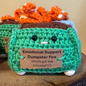 May include: A handmade, green crochet "Emotional Support Dumpster Fire" with black bead eyes and a white stitched mouth. The top features orange and white crochet flames. A brown tag reads "Who's got the s'mores!?!"