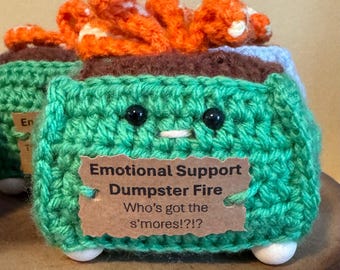 Peut inclure: Un "Emotional Support Dumpster Fire" au crochet vert fait à la main, avec des yeux en perles noires et une bouche cousue blanche. Le dessus présente des flammes au crochet orange et blanc. Une étiquette marron indique "Who's got the s'mores!?!"