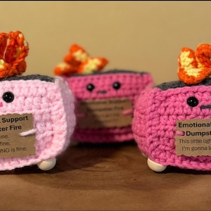 May include: Three handmade crochet "Emotional Support Dumpster Fire" figures. Each pink figure has a gray top, black eyes, and a small orange flame. One figure has the text "It's fine. I'm fine. EVERYTHING is fine."