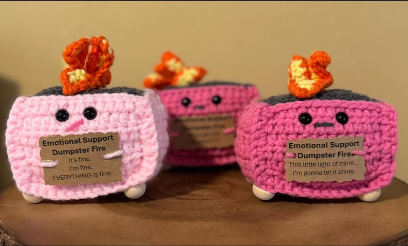 May include: Three handmade crochet "Emotional Support Dumpster Fire" figures. Each pink figure has a gray top, black eyes, and a small orange flame. One figure has the text "It's fine. I'm fine. EVERYTHING is fine."