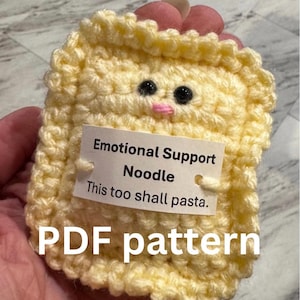 May include: A crocheted yellow noodle with black eyes and a pink mouth. A tag reads "Emotional Support Noodle. This too shall pasta." The words "PDF pattern" are also visible.
