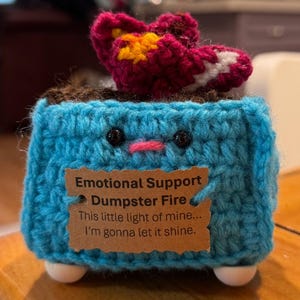May include: A handmade crochet sculpture of a blue dumpster with a smiling face, a red and yellow flame, and white feet. A brown tag reads "Emotional Support Dumpster Fire" with the quote "This little light of mine... I'm gonna let it shine."