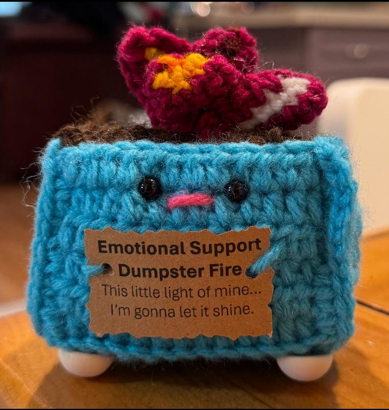 May include: A handmade crochet sculpture of a blue dumpster with a smiling face, a red and yellow flame, and white feet. A brown tag reads "Emotional Support Dumpster Fire" with the quote "This little light of mine... I'm gonna let it shine."