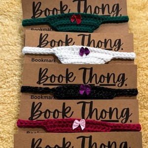 Può includere: Sei segnalibri all'uncinetto con il testo "Book Thong" su ciascuno. I segnalibri sono di diversi colori, tra cui nero, bianco, verde, rosso e viola. Ogni segnalibro ha un piccolo fiocco.