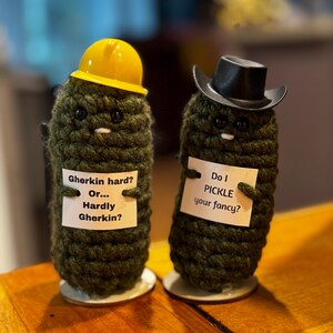 Könnte beinhalten: Zwei dunkelgrüne gehäkelte Gurkenfiguren auf runden Sockeln. Die linke Gurke trägt einen gelben Bauhelm und hält ein Schild mit der Aufschrift Gherkin hard? Or... Hardly Gherkin? Die rechte Gurke trägt einen schwarzen Cowboyhut und hält ein Schild mit der Aufschrift Do I PICKLE your fancy?