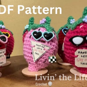 May include: Four crocheted strawberry amigurumi dolls wearing sunglasses and holding signs that say "Hang on! Let's try this..."  The dolls are pink and red and have green leaves.  The dolls are standing on small wooden bases.  The text "Livin' the Life" is written below the dolls.