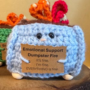May include: A light blue crochet dumpster fire with black eyes, a small beak, and wooden feet. The dumpster fire has orange and red flames. A brown tag reads "Emotional Support Dumpster Fire. It's fine. I'm fine. EVERYTHING is fine."