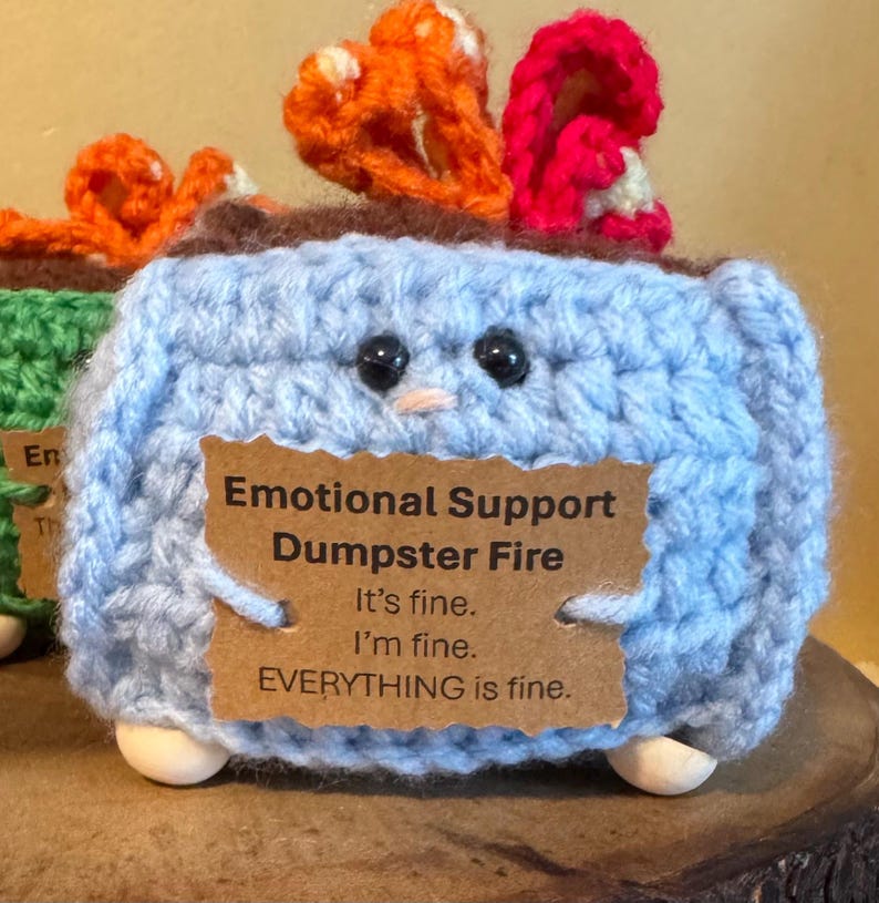 May include: A light blue crochet dumpster fire with black eyes, a small beak, and wooden feet. The dumpster fire has orange and red flames. A brown tag reads "Emotional Support Dumpster Fire. It's fine. I'm fine. EVERYTHING is fine."