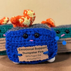 May include: Crocheted blue, green, and gray dumpster fire plush toys with orange and yellow flames. Each has black eyes, a pink mouth, and a tag that reads "Emotional Support Dumpster Fire".
